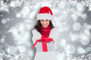 Festive brunette in santa hat giving gift  against shimmering light design on grey