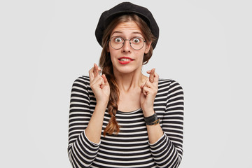 Shocked beautiful Parisian female crosses fingers as feels worried before exam, has desirable look, wears clothes in French style, isolated over white background. Gorgeous young Frenchwoman.