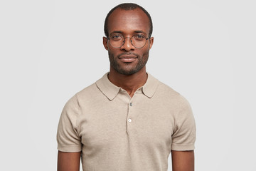 Fototapeta premium Confident young male manager with dark healthy skin, wears round spectacles and casual t shirt, looks seriously at camera, being sure in everything, has stubble, poses against white background