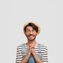 Vertical shot of happy male farmer makes praying gesture, asks God for good weather on tomorrow`s day as going to reap harvest, looks with great desire upwards, blank copy space for promotional text