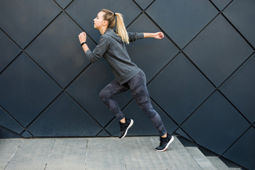 Young slim sports woman in fitness clothing jumping and running outdoors on wall background