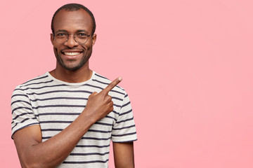 Positive black African American male seller advertises new products, rejoices discount, wears casual striped t shirt, isolated over pink background with blank space for your advertising content
