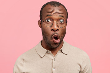 Candid shot of shocked dark skinned male model with bugged eyes and widely opened mouth, looks with surprisment at camera, poses against pink background. Surprised young African American man