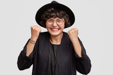 Overjoyed beautiful female hipster female clenches fists with happiness, has broad smile and delighted look, rejoices her promotion on work, wears stylish black hat and round glasses isolated on white
