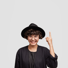 Lovely cheerful female architector demonstrates her work upwards, dressed in black long cloak, has pleasant smile. Satisfied woman in eyewear advertises product at blank copy space on studio wall