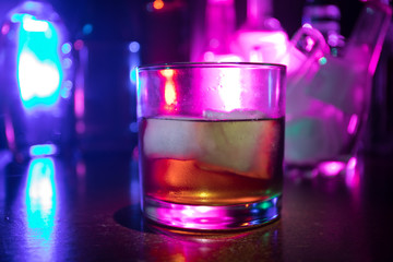 Glass of whisky on wooden bar closeup with bottles blurred view on dark background with light and smoke. Single glass of whisky on ice with a reflective wooden surface