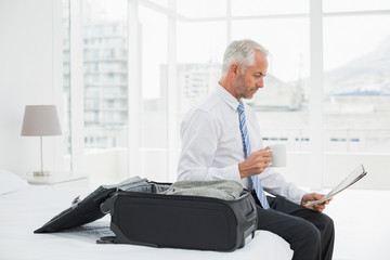 Businessman with coffee cup reading newspaper by luggage at hotel room