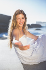 Fototapeta na wymiar Beautiful smiling blonde in sundress offering hand sitting on the beach
