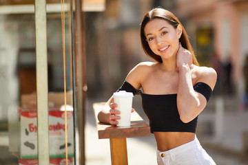 Brunette girl stand on the street look at camera and dring her delicious coffee from white cup, spring time photo shoot
