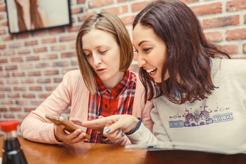 Two cheerful and beautiful girls are sitting together in cafe and texting sms by phone