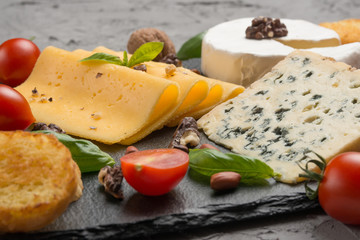 Different kinds of cheeses on a black board, with cherry tomatoes and basil and nuts.