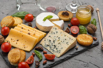 Different kinds of cheeses on a black board, with cherry tomatoes and basil and nuts.With red wine in a glass