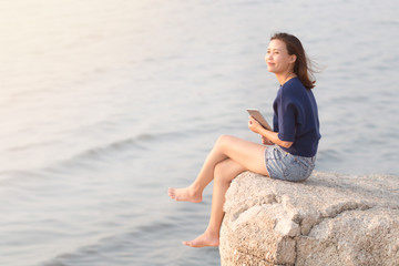 Fototapeta na wymiar Happiness young Asian woman relaxing and sitting on the rock cliff with smiling face and think about something good while holding or using digital tablet with blurred sea wave ocean background.