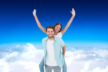 Fototapeta na wymiar Happy casual man giving pretty girlfriend piggy back against blue sky over clouds