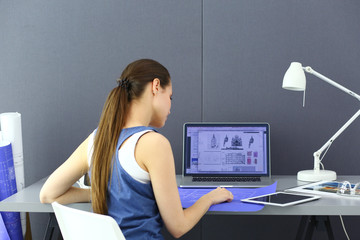 Young woman sitting at the desk with instruments, plan and laptop