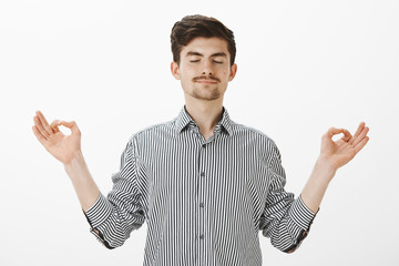 Naklejka premium Portrait of charming satisfied caucasian man with relaxed expression, spread hands in zen gesture, standing with closed eyes and pleased calm smile, meditating or practicing yoga over gray wall