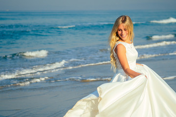 Fototapeta na wymiar young and beautiful bride with long blond hair smiling and walking on the ocean background in a beautiful white long wedding dress. a happy bride married a wedding ceremony on the beach by the sea