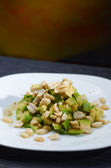 A simple salad of avocado and peanuts. Appetizer from vegetables, fruits, nuts and dressings. Food on a white plate and a black background. The appetizer is close-up.