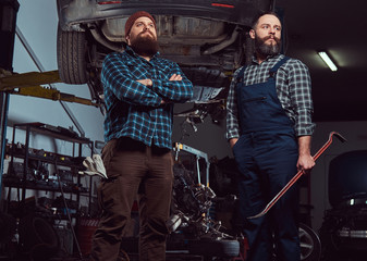 Two bearded brutal mechanics repair a car on a lift in the garage. 