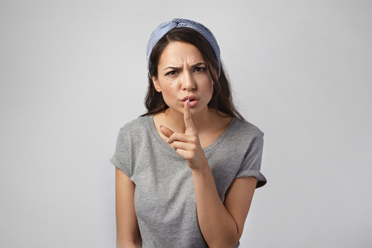 Picture of angry young brunette woman frowning, holding finger at her lips, saying Shh, asking to keep silence while she watching TV. Furious girl shushing at camera, telling not to make noise