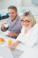 Couple in bathrobes with coffee and juice using laptop in kitchen