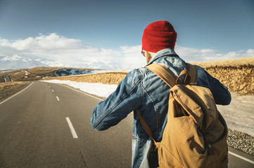 A bearded hipster with an old-fashioned vintage backpack wearing sunglasses with a red hat and jeans jacket and jeans stands on a countryside asphalt mountain road and looks at the snow-capped