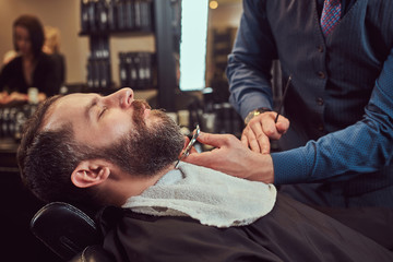 Professional hairdresser modeling beard with scissors and comb at the barbershop.