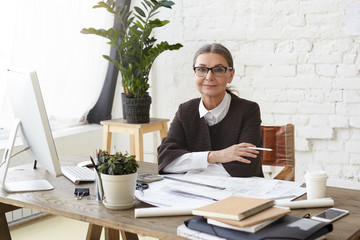 Attractive mature female architect in spectacles enjoying working process in light spacious office, sitting in front of generic computer, holding pencil, examining drawings and specifications on desk - Powered by Adobe