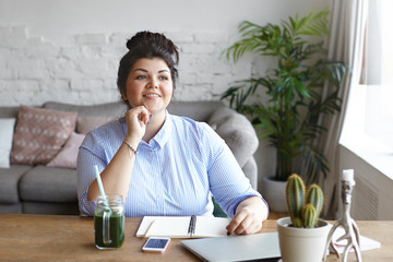 Horizontal shot of body positive chubby brunette woman writer with hair bun working at home with...