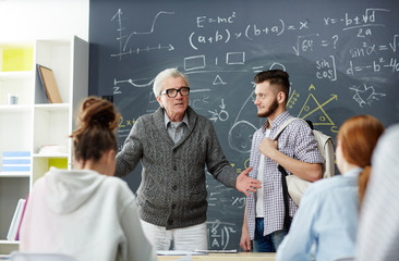 Mature teacher talking group of students about their new groupmate by blackboard at the beginning of lesson