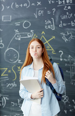 Teenage student with books and backpack blowing bubble gum by blackboard