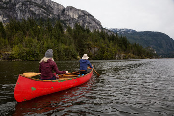 Adventurous people on a wooden canoe are paddling in a river with beautiful rocky mountain in the background. Taken in Squamish, North of Vancouver, BC, Canada.