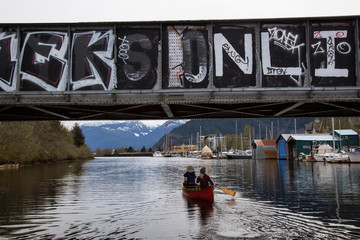 Fototapeta na wymiar Squamish, British Columbia, Canada - April 15, 2018: Couple friends are canoeing in the river near a train bridge.