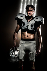 Fototapeta na wymiar Shirtless American football player with padding holding helmet against spotlights