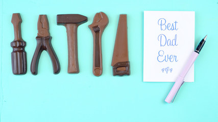 Happy Father's Day overhead of chocolate tool set with Best Dad Ever greeting card on pale blue aqua table.