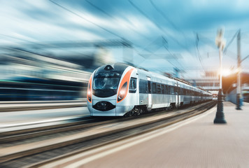 High speed passenger train in motion on the railway station at sunset in Europe. Modern intercity train on railway platform with motion blur effect. Urban scene with railroad. Railway transportation