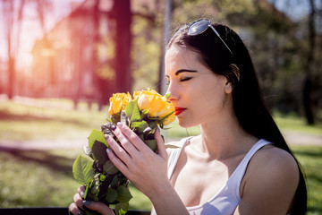 Attractive woman smelling the flowers. Satisfied and beautiful girl holds yellow roses in hands, closes eyes, sits on a park bench. Mom's day gift, birthday present, proposal.