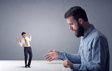 Young professional businessman being angry with an other miniature businessman in front of a grey...