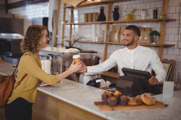 Fototapeta na wymiar Smiling waiter giving disposable coffee cup to young female customer at counter