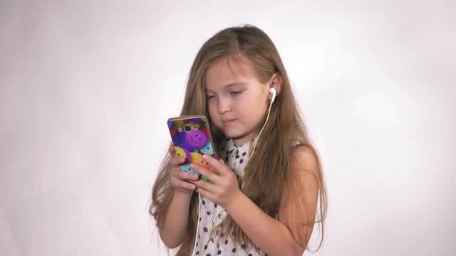 Child with earflaps dancing at studio background. The girl listens to music on the smartphone. The kid has loose long hair. Shooted on a gray white background.