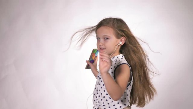 Child with earflaps dancing at studio background. The girl listens to music on the smartphone. The kid has loose long hair. Shooted on a gray white background.