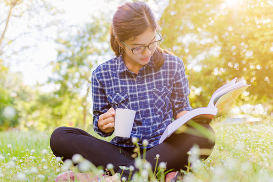 The girl sitting on a green grass with cup of coffee and reads the book