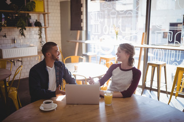 Smiling young friends talking while sitting with laptop at coffee shop
