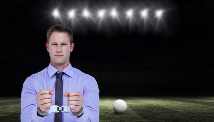 Handsome businessman wearing handcuffs against football pitch at night with ball and lights