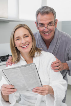 Happy couple in bathrobes reading newspaper in kitchen
