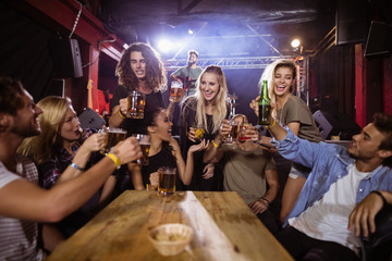 Cheerful friends toasting drink at table with performer singing on stage 