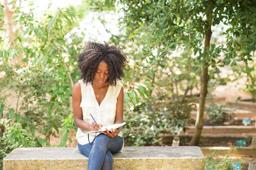 Focused attractive young black woman making notes in park. Girl sitting on concrete bench and...