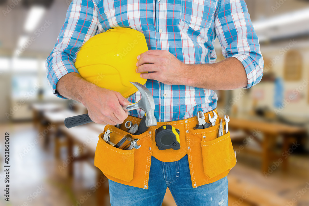Poster manual worker wearing tool belt while holding hammer and helmet against workshop