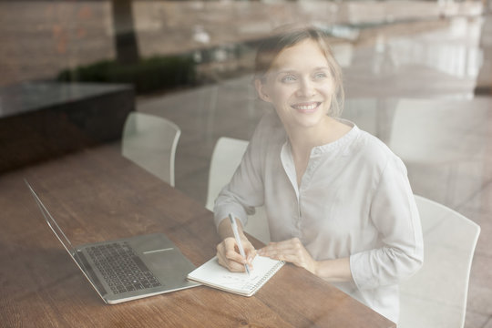 Smiling Beautiful Young Woman Making Notes And Working In Cafe. She Is Sitting At Table With Laptop Computer And Looking Away Through Window. Freelance Concept. View From Street Through Window Glass.