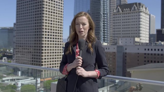 Medium shot, smartly dressed female news reporter talking with microphone in front of skyscrapers in the Financial District of Downtown Los Angeles. Hand-held, real time 4K UHD. Mute